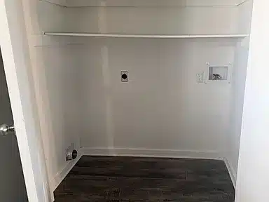 Empty laundry room with a dark hardwood floor, a shelf above, and hookups for a washer and dryer on a white wall.