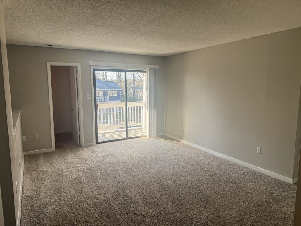 Empty carpeted room with beige walls, sliding glass door leading to a small balcony, and an open doorway on the left.