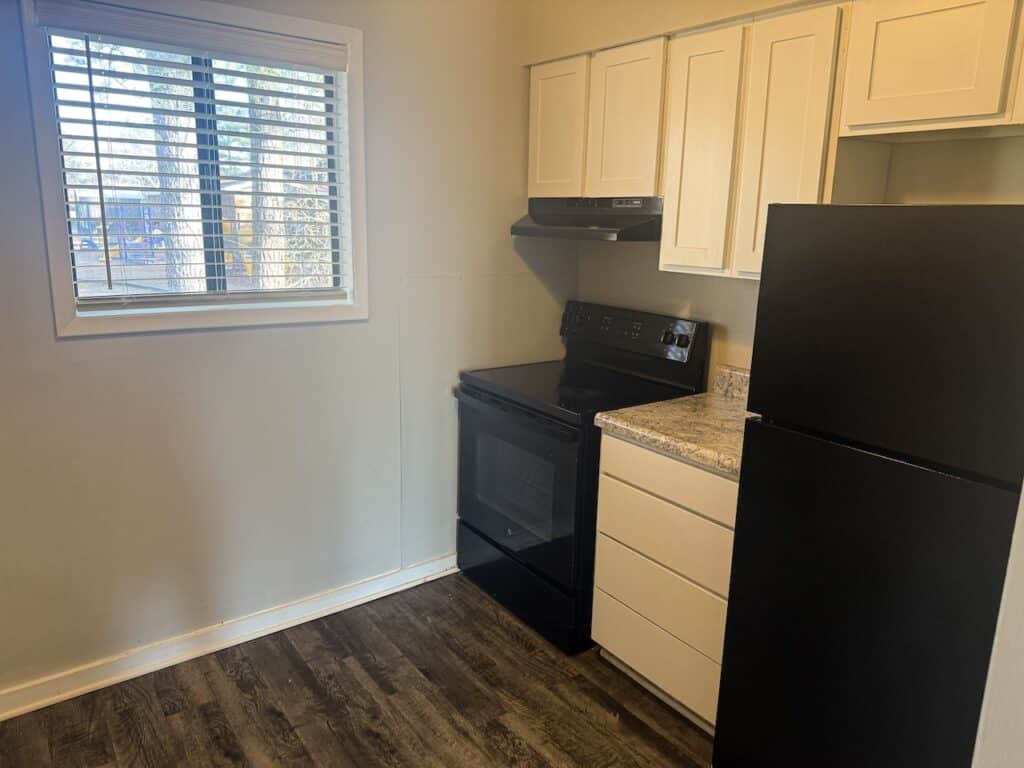 Small kitchen with a window, white cabinets, black appliances including a stove and refrigerator, and dark wood flooring.