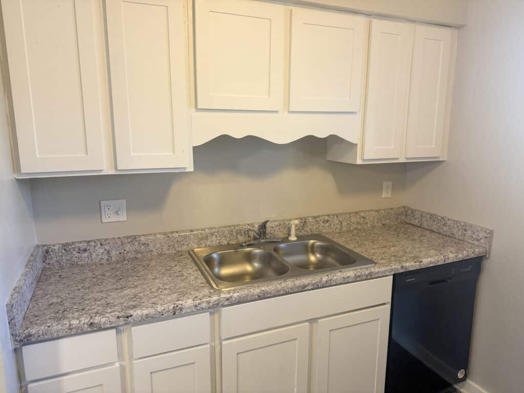 A kitchen with white cabinets, gray granite countertops, a stainless steel double sink, and a black dishwasher beneath the counter.