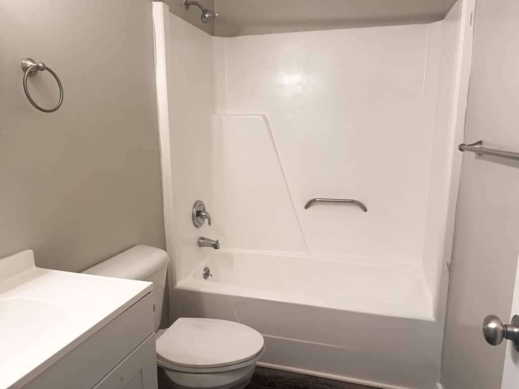 A bathroom with a white bathtub, shower fixtures, grab bar, toilet, and sink cabinet. Walls are light-colored, and hardware is metal.