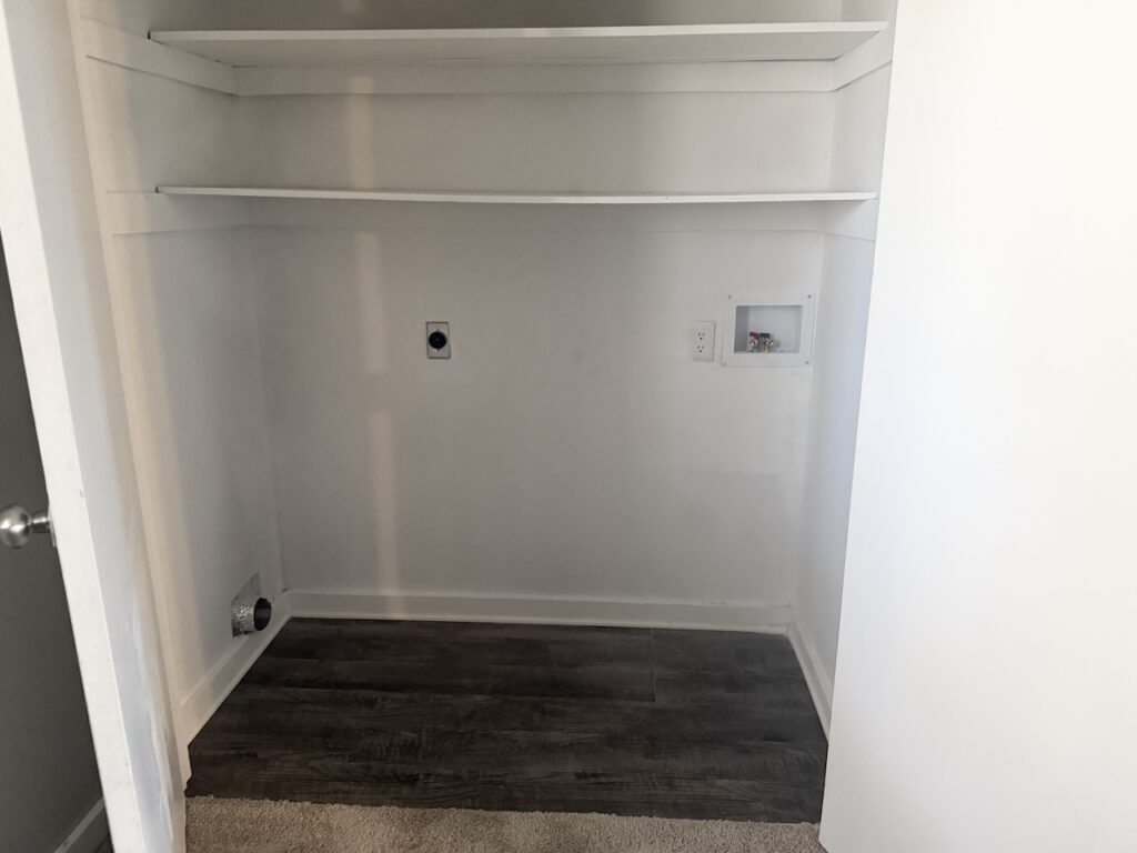 Empty utility closet with two shelves, a small recessed utility box, electrical outlets, and a circular vent on the left wall. Brown vinyl flooring and white walls.