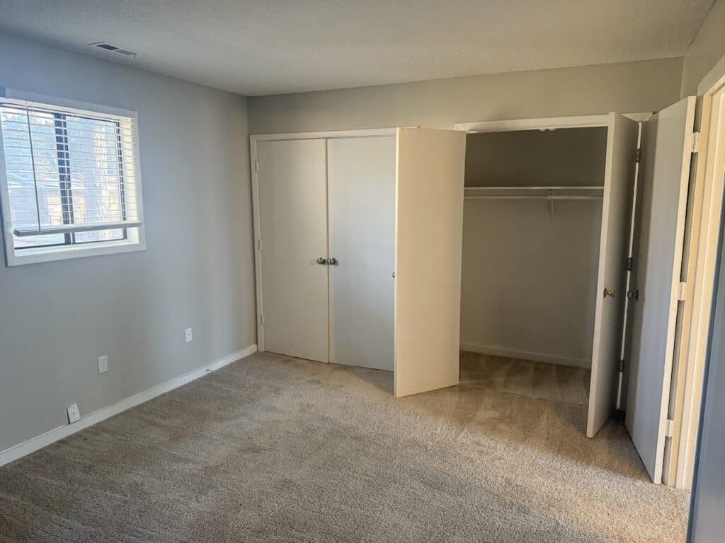 Empty carpeted room with beige walls, a window with blinds on the left, and an open closet with white double doors.