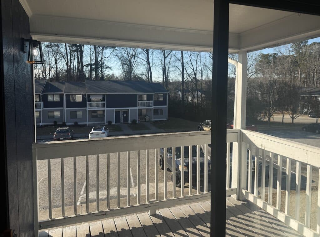 View from a second-story balcony showing a residential parking lot and a two-story building with cars parked outside. Bare trees and a clear sky are visible in the background.