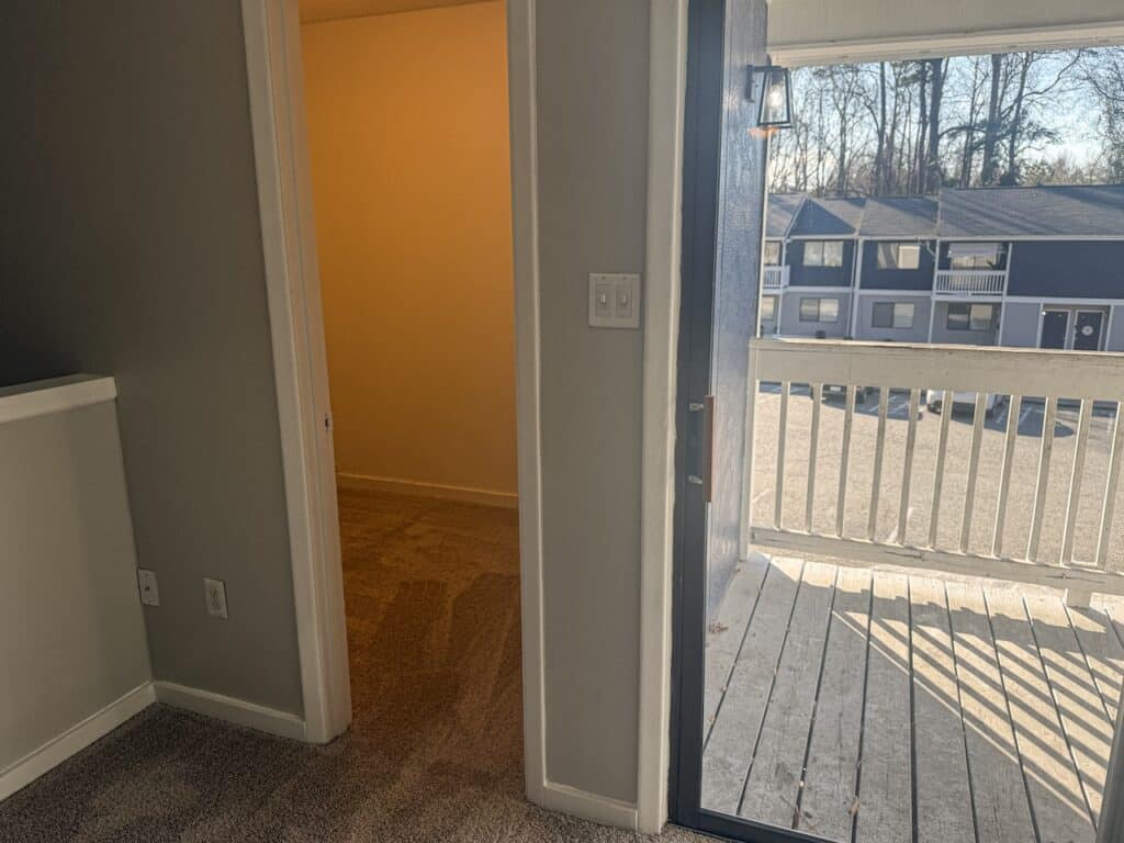 Interior view of a room with light gray walls and carpet, open to a balcony through a sliding door. The balcony has a wooden railing and overlooks a parking lot with cars.