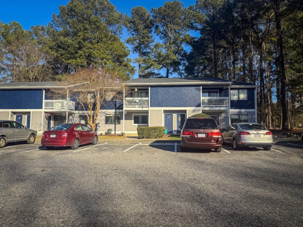 Apartment building with blue and gray exterior, surrounded by trees. Three cars are parked in front on an asphalt lot under a clear blue sky.