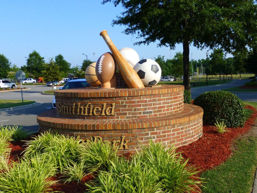 Brick monument with oversized sports equipment including a bat, ball, football, soccer ball, and volleyball. "Smithfield" is engraved on the brick. Surrounded by shrubs and trees.