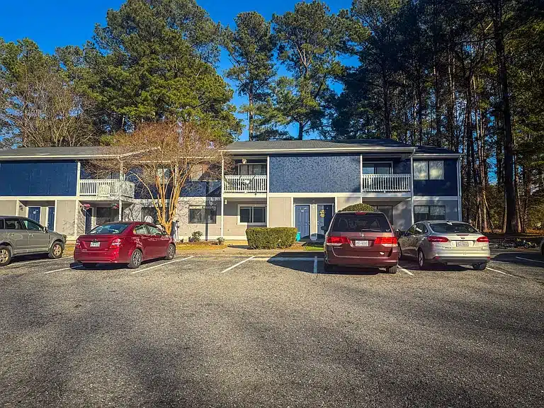 Apartment building with blue siding and white balconies in a wooded area. Four cars parked in the foreground on a sunny day.
