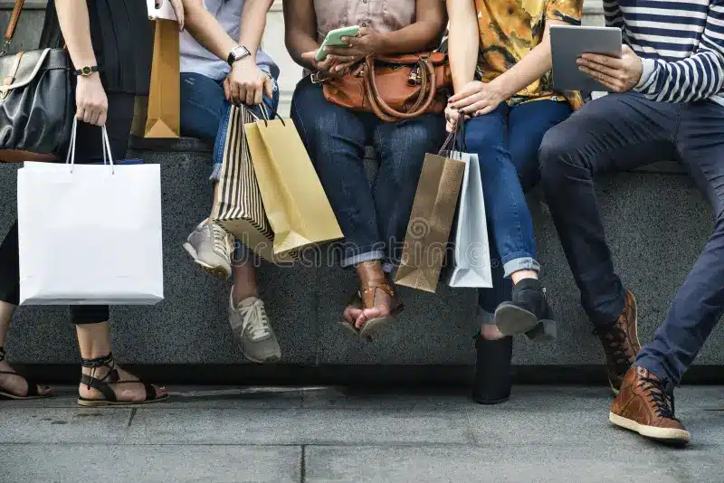 Five people sitting with shopping bags, using phones and a tablet, with legs and feet visible, wearing casual clothes and shoes on a concrete ledge.